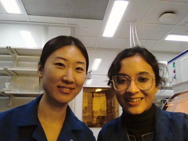Two smiling women in blue lab coats. One with dark hair pulled back, the other with glasses and dark curly hair, in a lab.
